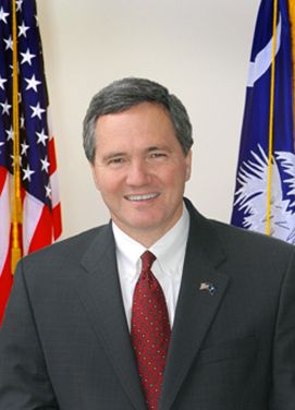 Speaker of the House of Representatives Bobby Harrell, smiling, standing between the United States of America flag and the state of South Carolina flag.